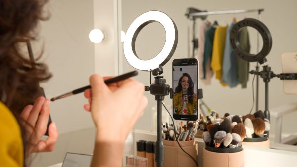 Glad Caucasian woman blogger girl influencer filming video mobile phone on stabilizer lamp studio female vlogger streamer talk demonstrating shadow palette do makeup tutorial beauty stream blog mirror